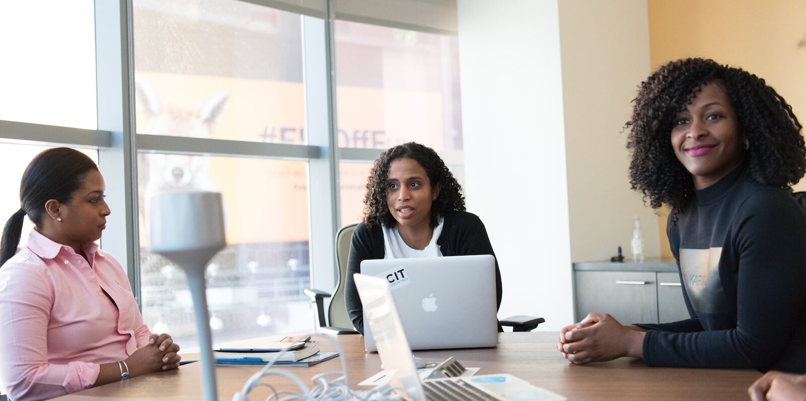 Three Woman Sitting Beside Brown Wooden Conference Table With Silver Apple Macbook